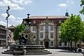 Mittig auf dem Erlanger Marktplatz steht der Paulibrunnen. Im Hintergrund bei blauem Himmel und leichter Bewölkung ist das Palais Stutterheim, indem die Stadtbibliothek und das Kunstpalais ihren Sitz haben, zu sehen.