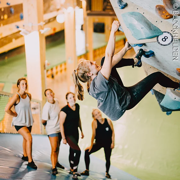 Blick auf eine Gruppe, die in der Boulderhalle klettert. Unter der Kletterwand liegt eine Schutzmatte.