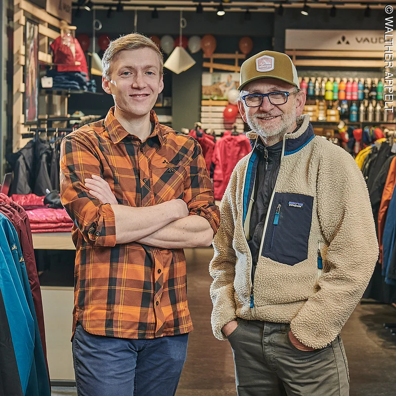 Herr Schmutzler mit Mitarbeiter im Verkaufsraum innen, im Hintergrund Jacken in vielen Farben, Trinkflaschen, Rücksäcke