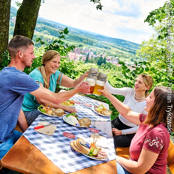 Auf einer mit blau-weiß karierten Tischdecke gedeckten Bierbank sitzt ein Mann und drei Frauen und stoßen mit einem Bier in der Hand an. Verschiedene Gerichte stehen auf dem Tisch. Im Hintergrund sieht man den tollen Blick ins Wiesenttal..