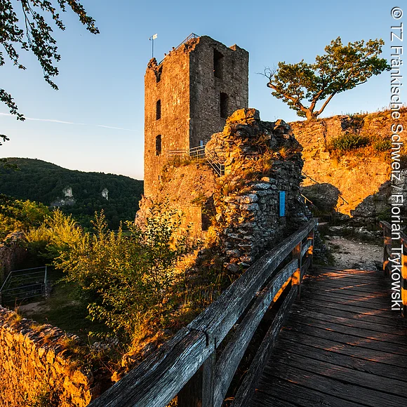 Eine alte Burgruine mit hohem Turm steht auf einem Felsen, im warmen Licht der untergehenden Sonne. Ein hölzerner Steg führt an den Mauerresten vorbei, umgeben von Bäumen und Hügeln.