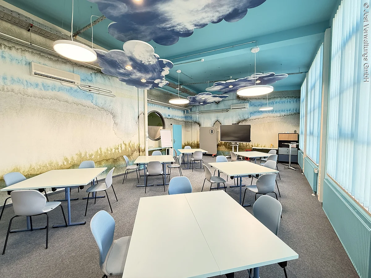 غرفة ندوات مع طاولات جماعية وعناصر تصميم هادئة باللون الأزرق والرمادي وسحب خفيفة على السقف.