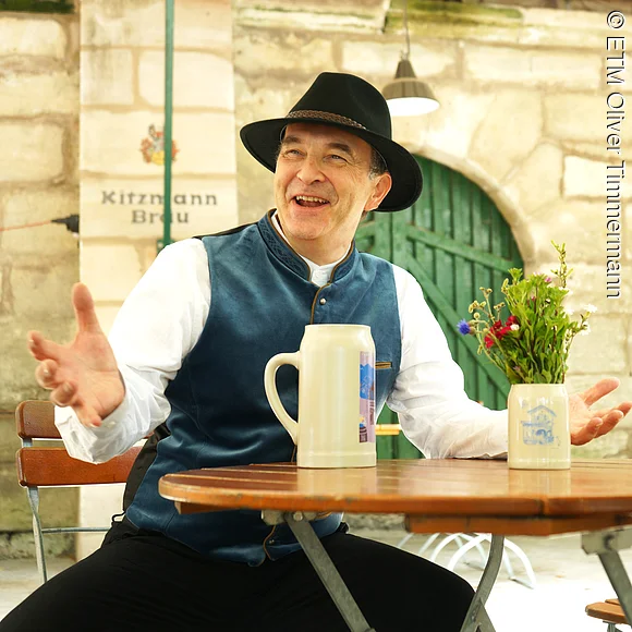 Wir sehen den kostümierten Stadtführer als Bierkutscher Schorsch, sitzend vor dem Kitzmann Keller in Erlangen, stilecht mit einem Ton-Bierkrug.