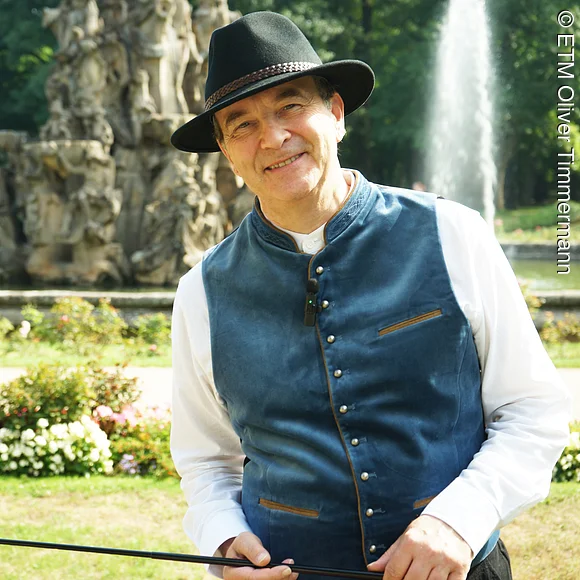 Wir sehen einen kostümierten Stadtführer als Bierkutscher Schorsch, stehend vor dem Hugenottenbrunnen im Schlossgarten mit einer Peitsche in der Hand.