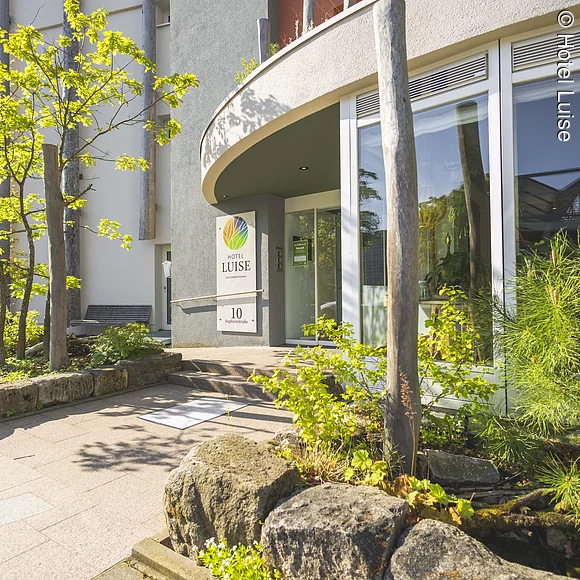 Hoteleingang mit großen Fenstern und außen vielen Pflanzen